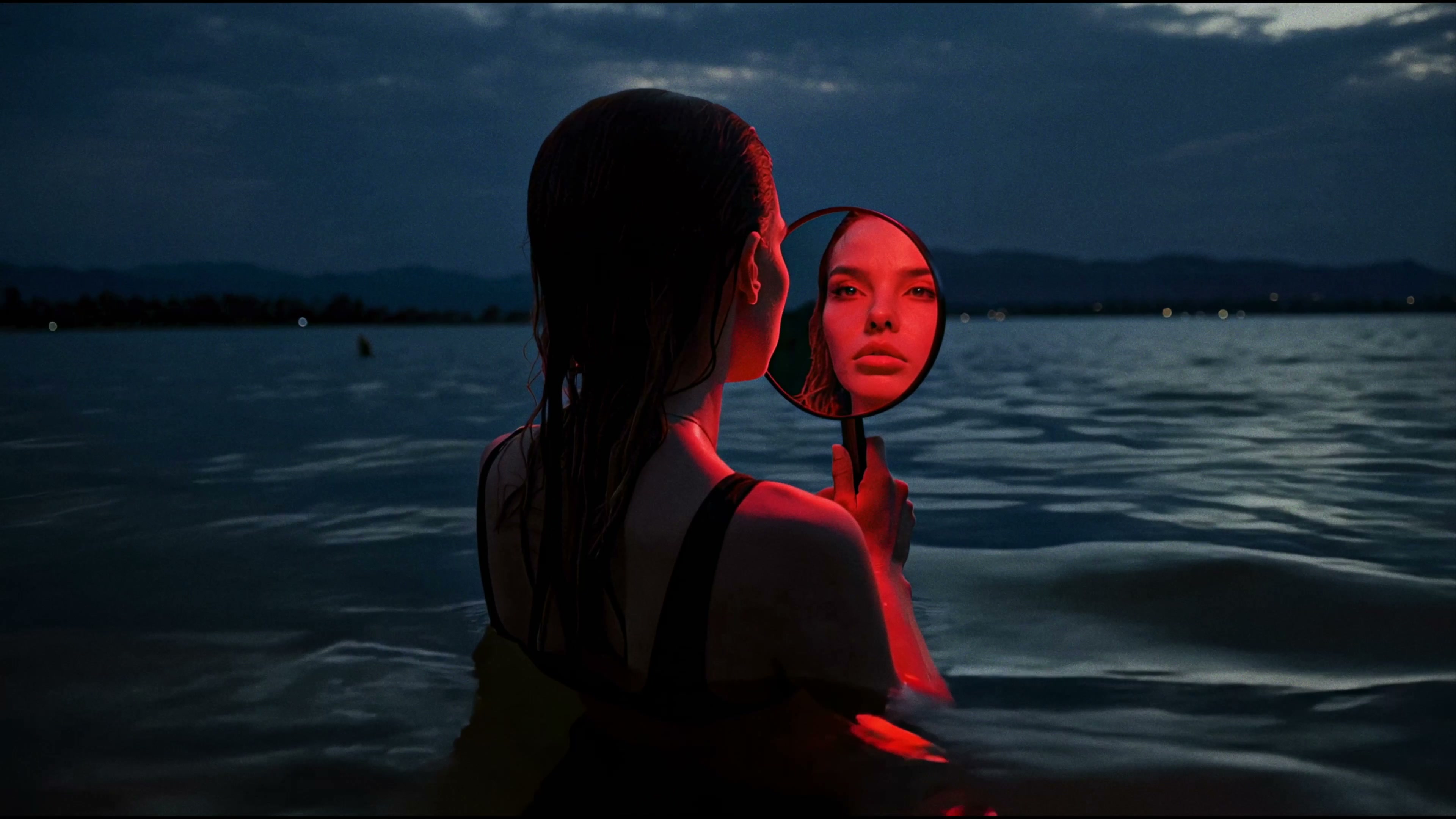 Alexandra Oz. in «Echoes» — standing in dark water at dusk holding a round hand-mirror, her reflection lit red.